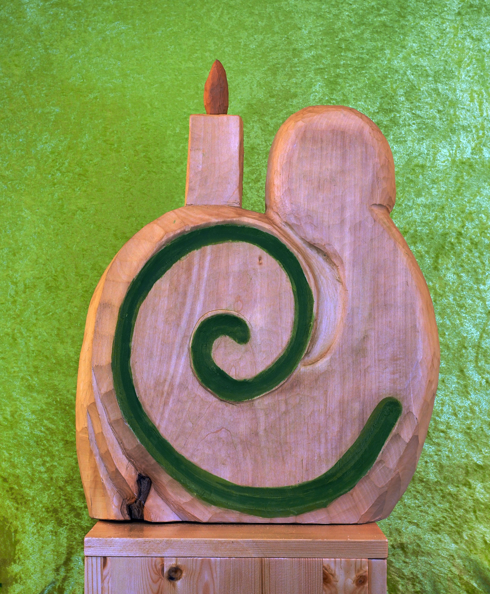 Holzskulptur mit einem stilisierten Schneckendesign. Das Schneckenhaus weist eine markante grüne Spirale auf einer hellen Holzoberfläche auf. Die Gesamtform ist abstrakt, mit einem grünen Wirbel und einer abgerundeten Gestalt. Die Skulptur steht vor einem leuchtend grünen, strukturierten Hintergrund und ruht auf einem Holzsockel.
