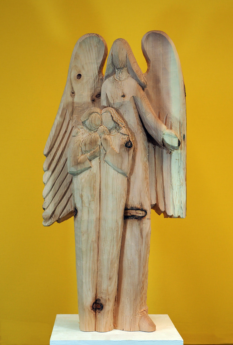Holzskulptur eines Engels mit Flügeln, auf einem Sockel vor einem gelben Hintergrund stehend. Der Engel hält eine Figur eines Kindes vor sich. Das Holz zeigt natürliche Astlöcher und Texturen.
